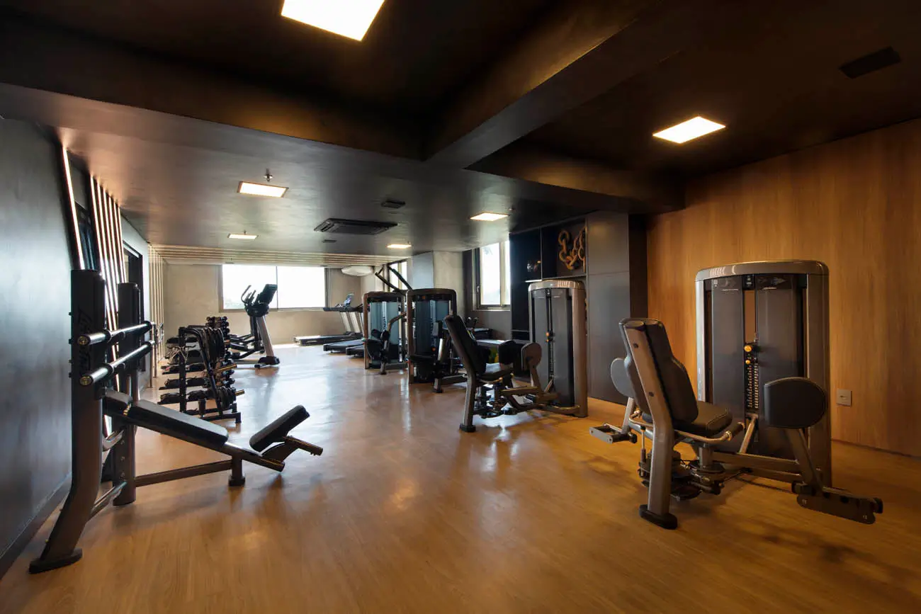 Academia com aparelhos de musculação, bancos de treino e janelas ao fundo.