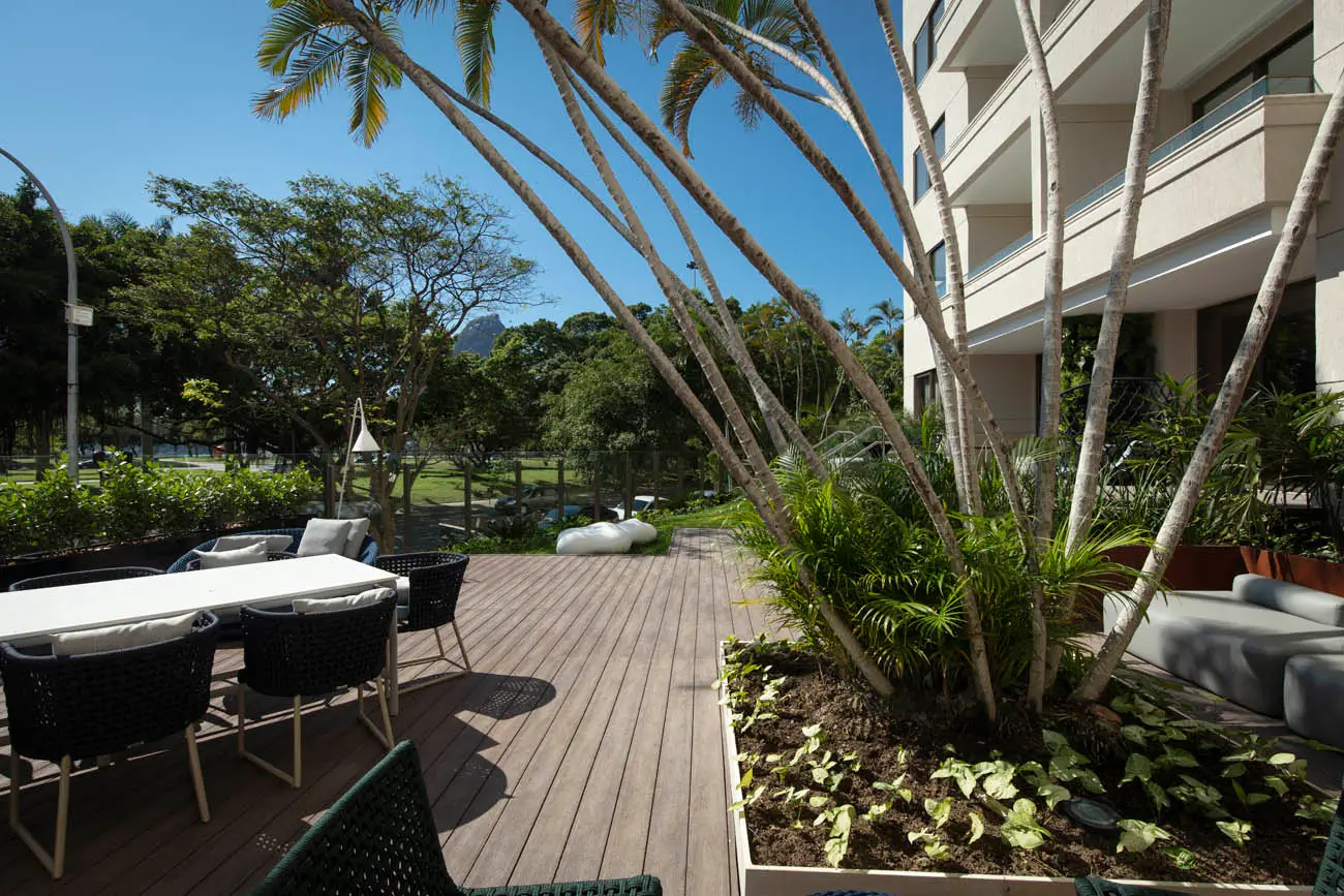 Deck de madeira com mesa e cadeiras escuras, palmeiras e fachada com varandas à direita.