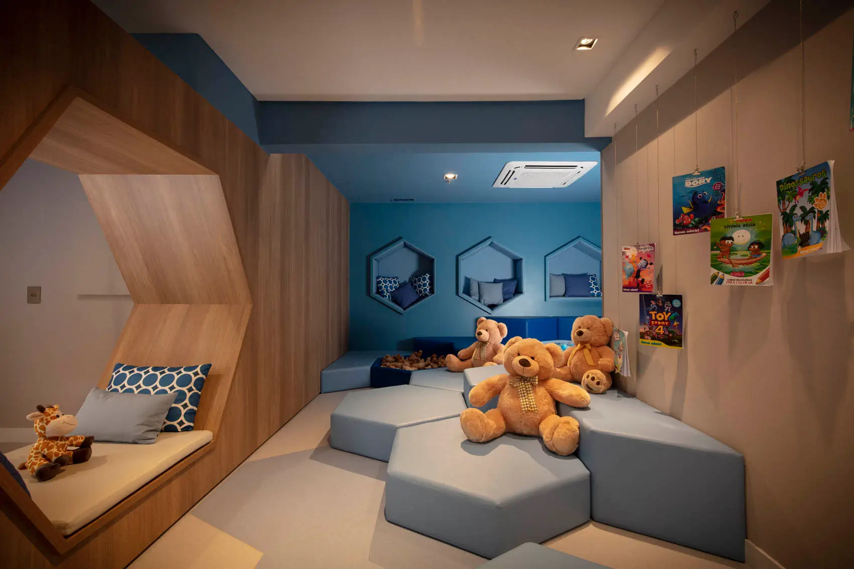 Brinquedoteca com módulos estofados cinza, ursos de pelúcia, nichos em forma de casa e parede azul.