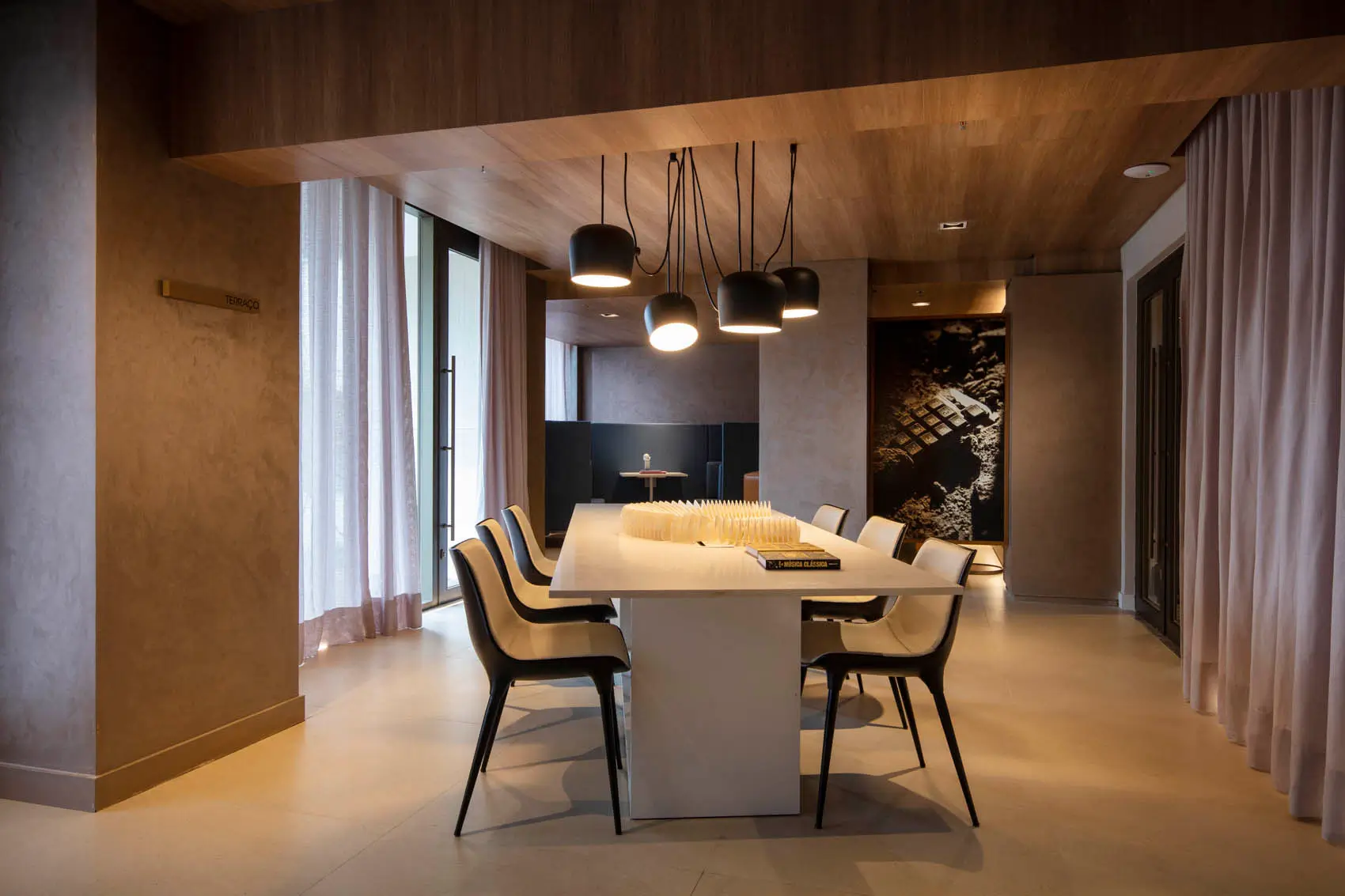 Coworking com mesa branca comprida, cadeiras claras, luminárias pendentes e revestimento de madeira.