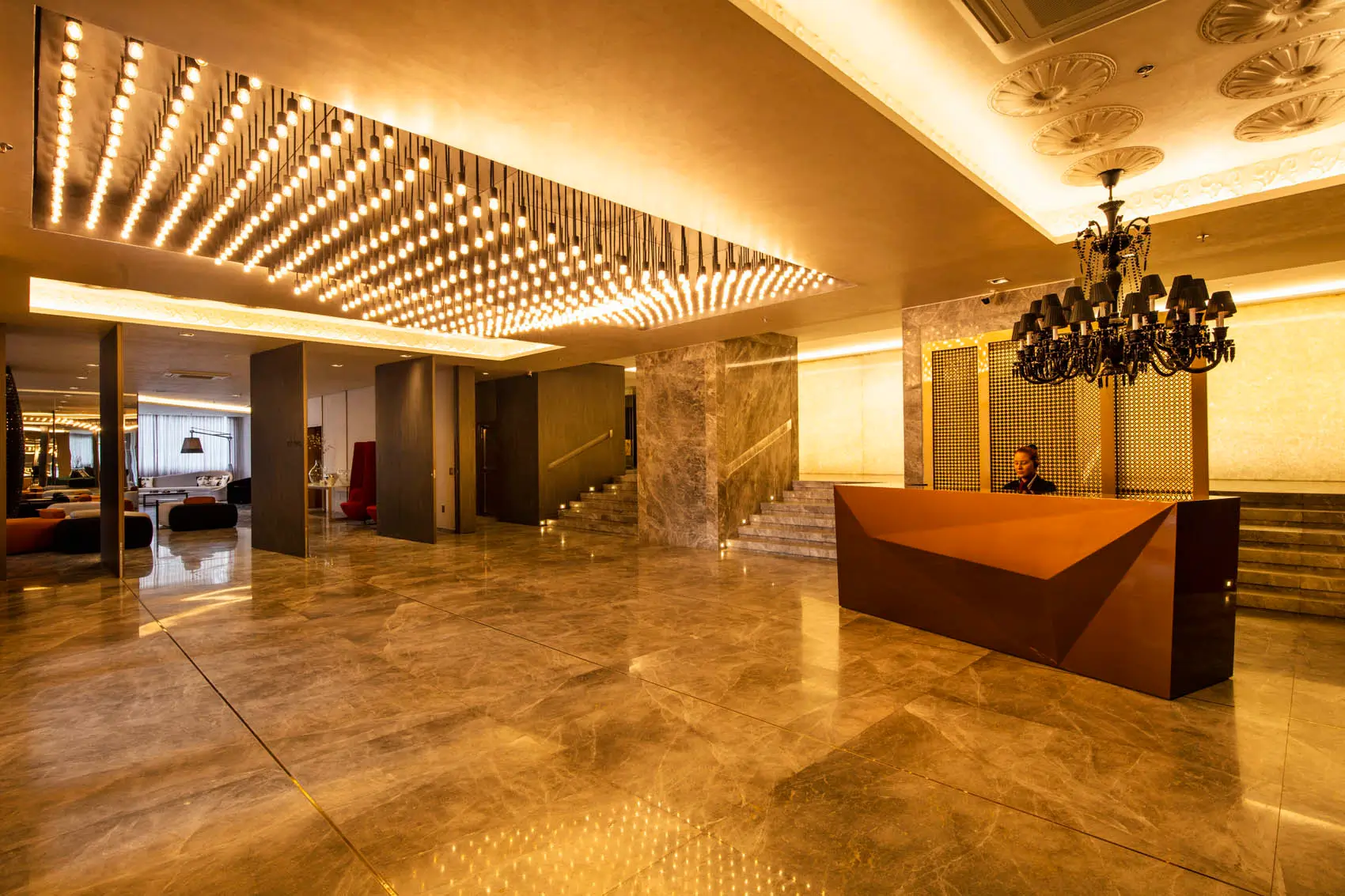 Lobby com balcão de recepção angular, lustres e numerosas lâmpadas pendentes sobre o piso de pedra.