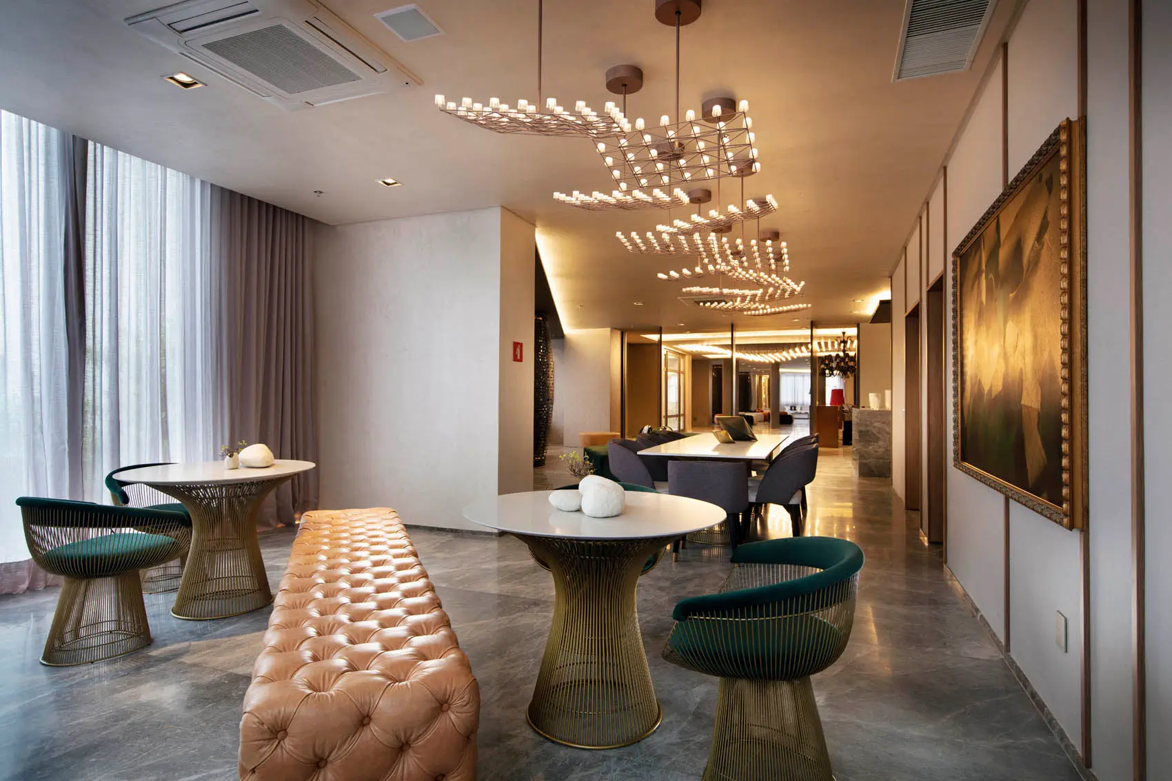 Lounge com mesas redondas, cadeiras verdes, banco de couro e luminárias geométricas suspensas.
