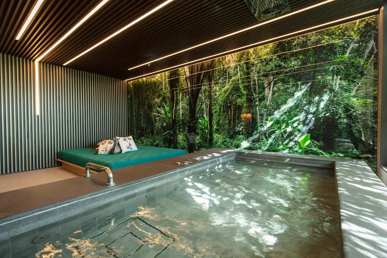 Hidromassagem retangular diante de uma parede envidraçada com vegetação, ao lado de um divã verde.