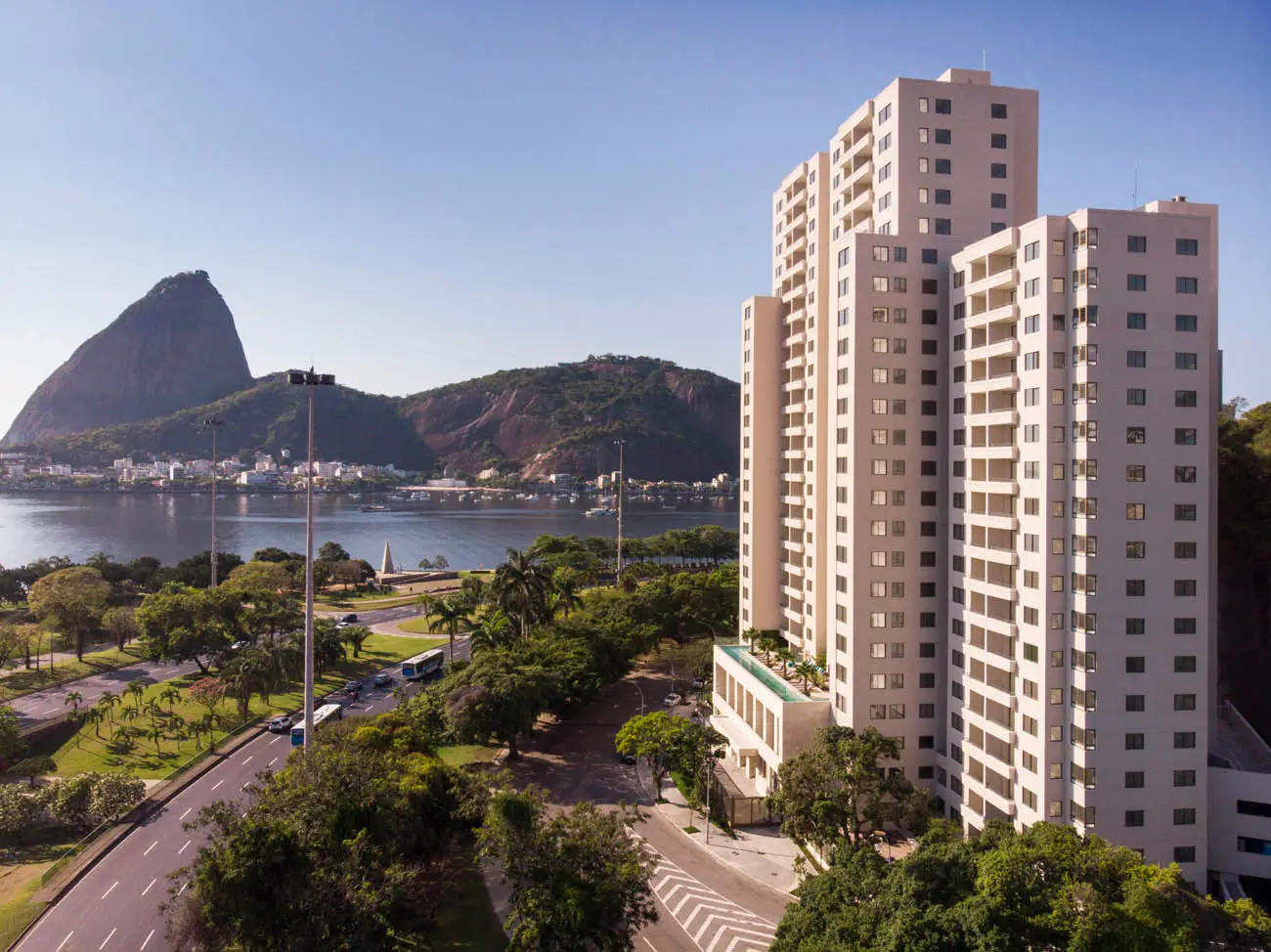 Vista externa das torres claras do Rio By Yoo, com avenida arborizada, água e Pão de Açúcar ao fundo.