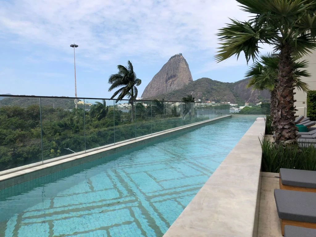Piscina alongada junto a um guarda-corpo de vidro, com palmeira à direita e Pão de Açúcar ao fundo.