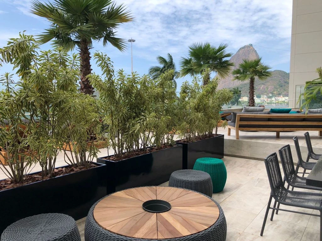 Terraço com mesa redonda de madeira, pufes verdes e cinza, jardineiras e Pão de Açúcar ao fundo.