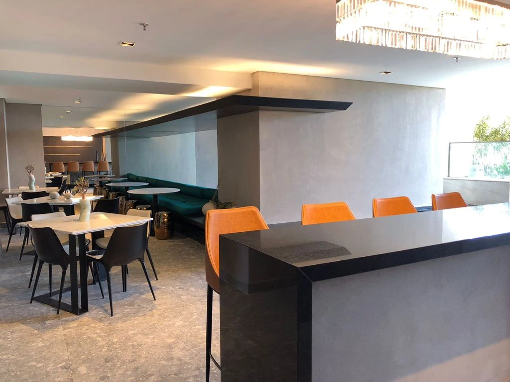 Bancada escura com banquetas laranja em primeiro plano e mesas com cadeiras ao fundo.
