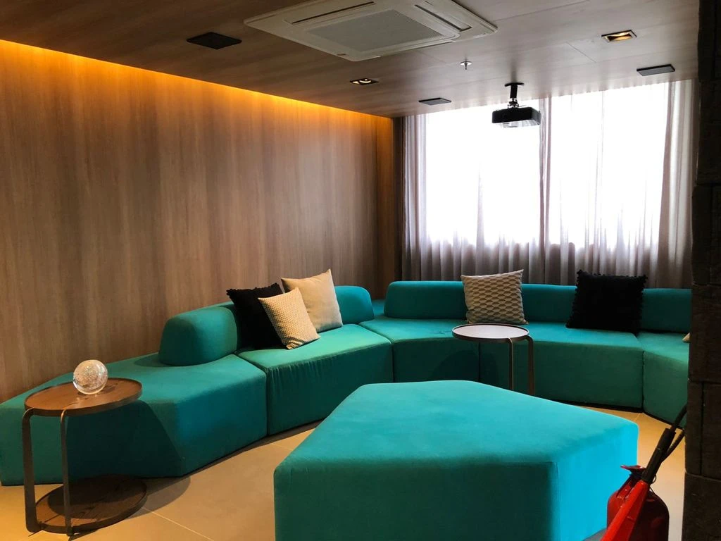Sofá modular verde em formato de U, pufe central e projetor no teto diante de uma janela com cortina.