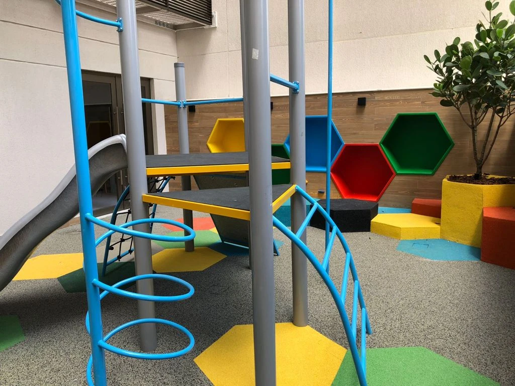 Playground com escorregador, estrutura de escalada azul, piso colorido e nichos hexagonais na parede.