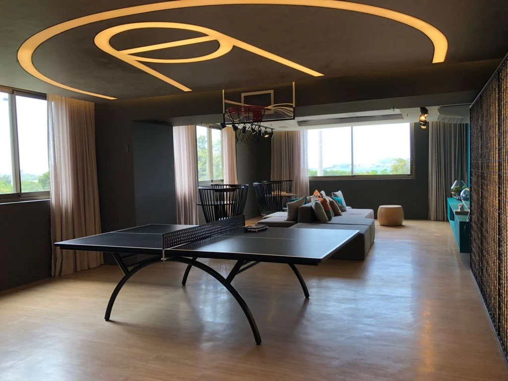 Sala com mesa de tênis de mesa, cesta de basquete, sofá ao fundo e iluminação curva no teto.