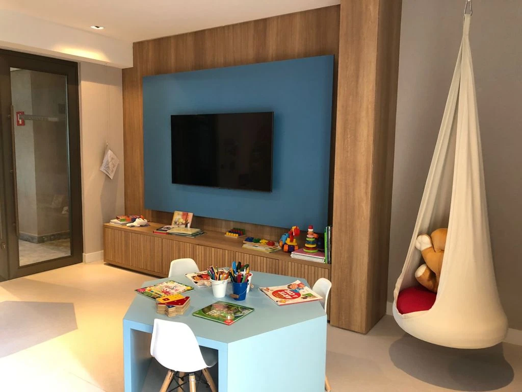 Mesa infantil azul com livros e lápis, cadeiras brancas, televisão na parede e assento suspenso de tecido.