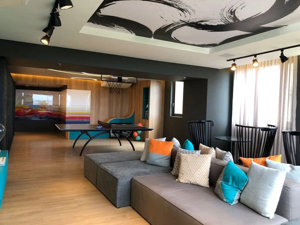 Sofá modular cinza com almofadas coloridas, mesa de tênis de mesa ao fundo e grafismo preto e branco no teto.