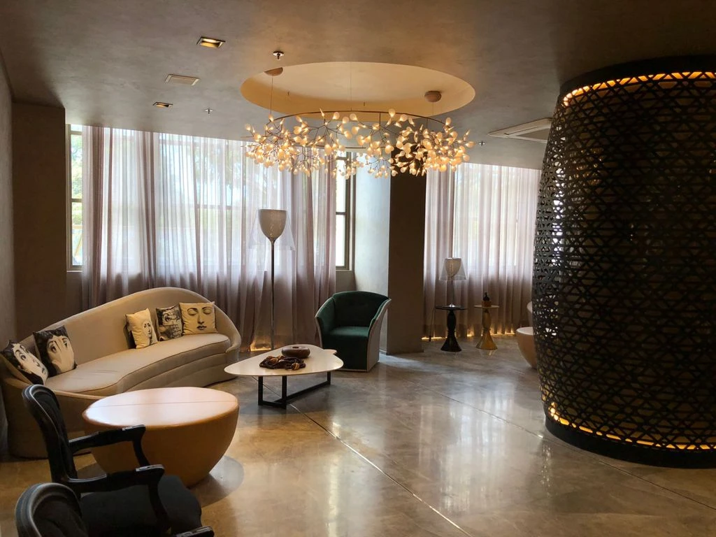 Lounge com sofá claro curvo, poltronas, lustre ramificado e coluna escura revestida por trama iluminada.